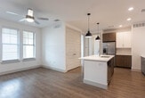 This single-floor residence at 1301 E Mockingbird Ln in Midland, Michigan offers a thoughtfully designed layout with modern finishes throughout. The kitchen serves as the heart of the space, featuring a large center island with a white quartz countertop and undermount sink, two stylish black pendant lights, and a two-tone cabinetry scheme pairing crisp white upper cabinets with warm wood-toned lower cabinets. A classic subway tile backsplash complements the stainless steel appliances, including a French door refrigerator, range, and built-in microwave. Warm-toned wood-look flooring runs consistently throughout the entire unit, creating a cohesive and inviting atmosphere. The open living area adjoins the kitchen and benefits from natural light through multiple windows fitted with horizontal blinds, along with a ceiling fan overhead. The bedroom is a bright, clean space with a large window offering exterior views and generous proportions. A spacious walk-in closet with a full-length hanging rod and shelf provides ample storage. The bathroom is well-appointed with a wood-toned vanity, white quartz countertop, a tub-shower combination with white tile surround, and brushed nickel fixtures. The unit's clean white walls and recessed lighting contribute to a bright, contemporary ambiance throughout.