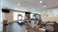 This single-floor fitness center is designed to support a full range of workout routines in a functional and energizing environment. The space is organized into distinct cardio and strength training zones. The cardio area houses a row of treadmills and elliptical trainers positioned along large windows fitted with horizontal blinds, allowing ample natural light to fill the room. Wall-mounted flat-screen televisions are positioned for convenient viewing during workouts. Full-length mirrors line one wall, visually expanding the space and allowing users to monitor their form.

The strength training zone includes cable machines, a multi-use weight bench, a dumbbell rack, and medicine balls, providing a comprehensive selection of resistance equipment. A bold red accent wall serves as a visual focal point in this area, complemented by a large motivational typography mural featuring words such as "inspire," "strength," "motivation," and "commit" in varying fonts and sizes, contributing to an encouraging atmosphere.

The flooring throughout is dark rubber, offering durability and cushioning suited for high-impact activity. Recessed lighting and flush-mount ceiling fixtures provide consistent, even illumination across the space. The overall design is practical and purposeful, balancing accessibility with a motivating aesthetic.