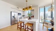 Located along First Avenue South in the heart of downtown Saint Petersburg, Florida, this single-floor residence delivers a refined urban living experience defined by clean contemporary design and abundant natural light. Large floor-to-ceiling windows are a defining architectural feature throughout, flooding the interior with sunlight and framing impressive views of the Saint Petersburg skyline.

The open-concept kitchen is outfitted with white cabinetry, quartz countertops, stainless steel appliances, and a central island with bar seating. A striking gold multi-pendant chandelier anchors the space with a warm, elevated aesthetic. Light hardwood flooring extends throughout the main living areas, lending a cohesive and airy feel.

The dining area benefits directly from the dramatic window line, offering panoramic city views that serve as a compelling backdrop. The living room is calm and well-proportioned, with recessed lighting and easy access to the surrounding rooms.

The bedrooms feature plush carpeting and generous walk-in closet space with built-in shelving. The primary bathroom is appointed with a glass-enclosed walk-in shower, white tile, quartz vanity surfaces, and gold hardware accents. A clean, well-lit entryway with light-toned walls completes the thoughtfully designed layout.