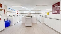 Located at 30 Godstone Rd in Toronto, Ontario, this communal laundry facility offers a clean, efficiently organized space designed to serve building residents. The room is finished with light-toned large-format floor tiles and white wall tiles accented by a distinctive gold horizontal band, lending a tidy and cohesive aesthetic throughout.

Rows of white top-loading washing machines line both sides of the room, with additional units positioned centrally, providing ample capacity for residents. A standalone dryer is also centrally placed, complementing the generous number of washing machines. Overhead, bright fluorescent lighting ensures excellent visibility across the entire space.

A built-in folding counter with a dark laminate surface runs along one wall, offering residents a practical area for sorting and folding laundry. A second stainless steel folding counter is mounted along another wall, adding further functionality. A metal bench near the entrance provides a comfortable place to wait.

The walls display posted notices and resident guidelines, and a fire extinguisher is visibly mounted for safety compliance. A bulletin board near the entry door keeps residents informed. Small windows along the upper portion of the walls allow for natural light and ventilation. The overall ambiance is practical, well-maintained, and designed to accommodate the day-to-day laundry needs of the building's residents efficiently.