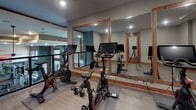 Located at 1121 3rd Ave N in Nashville, Tennessee, this single-floor fitness studio is thoughtfully designed to support a range of exercise activities. The space is finished with light wood-tone luxury vinyl flooring throughout, complemented by warm wood baseboards and trim that lend a polished, cohesive aesthetic to the interior.

The main workout area is equipped with multiple Peloton stationary bikes, each outfitted with digital display screens, and is lined with large wood-framed mirrors that visually expand the room and allow users to monitor their form during exercise. Wall-mounted ballet barres run beneath the mirrors, adding versatility for stretching, barre workouts, and strength training. Exercise balls and camera equipment are also present, supporting both in-person and recorded fitness sessions.

An adjoining open room provides flexible, multipurpose space that can be configured for additional training or storage needs. Glass-paneled doors with wood frames separate zones within the studio while maintaining an open, airy feel. Recessed lighting throughout keeps the environment well-lit and functional. The overall design balances clean, modern finishes with warm wood accents, creating a professional yet welcoming atmosphere suited for focused fitness use.