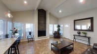 This single-story clubhouse offers an inviting and well-appointed gathering space designed to impress. The centerpiece of the main room is a dramatic floor-to-ceiling fireplace clad in dark wood shiplap, rising to meet the soaring vaulted ceiling and creating a bold, striking focal point. Exposed ceiling beams add architectural character and reinforce the grand sense of height throughout the space. The open layout is bathed in natural light, courtesy of multiple large windows fitted with white blinds that allow for flexible light control. Warm-toned ceramic tile flooring extends throughout, providing both durability and a cohesive aesthetic. The room is thoughtfully furnished with a substantial wooden coffee table, a classic leather armchair, and decorative accent seating, creating a comfortable and functional environment for residents to gather. A dedicated entryway with an arched transom window provides a welcoming first impression upon arrival. Recessed lighting complements the natural light, ensuring the space is well-illuminated at all hours. The overall ambiance balances casual comfort with refined design, making this clubhouse a versatile and attractive amenity for any community.