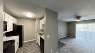 Located at 7643 E 51st St in Tulsa, Oklahoma, this single-floor unit offers a clean, modern aesthetic throughout. The galley-style kitchen is a standout feature, equipped with white shaker cabinets, granite-look countertops, black appliances including a refrigerator, range, and dishwasher, and a stainless steel sink. Dark wood-look vinyl plank flooring runs through the kitchen, providing a sharp contrast to the neutral gray walls and white trim. The living area flows naturally from the kitchen and is finished with plush gray carpet underfoot. A ceiling fan overhead and a large sliding glass door with vertical blinds allow for both airflow and an influx of natural light, giving the living space an open, airy feel. The front entry door is also located within this main living space, keeping the layout straightforward and functional. The bathroom is well-finished, featuring a granite-top vanity with a white undermount sink, white cabinetry with modern hardware, a lighted vanity mirror, a bathtub with tiled surround, and wood-look flooring that ties back to the kitchen aesthetic. A dedicated laundry connection is also present. Throughout the unit, a consistent warm taupe color palette and textured ceilings create a cohesive and inviting atmosphere.