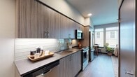 This thoughtfully designed single-floor studio offers a cohesive open-plan layout that seamlessly integrates the living, sleeping, and dining areas into one bright, functional space. Large windows with slatted blinds allow generous natural light to fill the room while framing views of the surrounding building exterior, contributing to an airy and welcoming ambiance throughout.

The galley-style kitchen is a standout feature, outfitted with floor-to-ceiling woodgrain cabinetry in a contemporary charcoal tone, sleek quartz-style countertops, and a glossy subway tile backsplash with under-cabinet lighting. Stainless steel appliances — including a full-size refrigerator, electric range, over-the-range microwave, and dishwasher — provide a complete and modern cooking experience.

The main living area accommodates a full-size bed alongside a cozy seating arrangement with a sofa, accent chairs, and a coffee table, all anchored by a patterned area rug over warm wood-tone flooring. A walk-in closet provides practical storage directly off the sleeping zone, while an additional closet is accessible from a separate corridor.

The bathroom features a clean, neutral palette with white subway tile surrounding a full tub-shower combination, complemented by hardwood-style flooring and simple, elegant fixtures. Throughout the space, the design maintains a consistent modern aesthetic with soft neutral tones, quality finishes, and practical yet stylish furnishings.