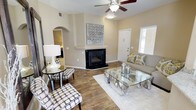 This single-floor home offers a thoughtfully designed living space that balances comfort and style. The main living area serves as the heart of the home, anchored by a striking corner fireplace clad in dark tile with a clean white mantel surround — a standout architectural feature that adds warmth and character to the room. Rich hardwood-style flooring runs throughout, providing a durable and elegant foundation that ties the space together seamlessly. Natural light filters in through a window fitted with blinds, brightening the neutral, warm-toned walls that create an inviting and relaxed atmosphere. A ceiling fan with an integrated light fixture ensures year-round comfort and adds a functional overhead element to the room. A distinctive arched doorway leads to an adjacent area, lending an element of architectural charm and providing a graceful transition between spaces. The layout is open and easy to navigate, making the most of the available footprint. Overall, the space presents a clean, move-in-ready aesthetic with classic design details, practical features, and a cohesive color palette that appeals to a wide range of tastes.