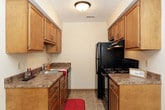 This single-floor residence offers a practical and well-organized layout across several distinct living areas. The kitchen is equipped with warm honey-toned wood cabinetry, granite-look laminate countertops, a double stainless steel sink, and a suite of black appliances including a refrigerator, dishwasher, and range. Light-colored tile flooring completes the functional cooking space.

The living area is generously proportioned, featuring neutral carpeting, a textured ceiling, and a ceiling fan with integrated lighting. A sliding glass door with vertical blinds allows natural light to fill the space while providing access to an outdoor area. Additional windows fitted with vertical blinds further brighten the room.

Multiple bedrooms are present throughout the unit, each finished with soft neutral carpeting, wood-trimmed doors, and ceiling fans. Closet space is well-represented, with louvered bifold and standard hinged closet doors visible across the rooms. The primary bedroom includes a notably spacious layout.

The bathroom features a wide vanity with a white countertop, wood cabinetry beneath, a large mirror, and classic small-tile flooring in a neutral tone. Overall, the space maintains a consistent, understated aesthetic with warm wood accents and light neutral walls throughout.