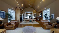 Located at 1000 Bally Bunion Drive in Egg Harbor, NJ, this single-story clubhouse offers a warm and well-appointed gathering space designed with both comfort and elegance in mind. The centerpiece of the interior is a grand open lounge area distinguished by soaring vaulted ceilings adorned with decorative string lighting and a ceiling fan, creating an airy yet intimate atmosphere. Rich hardwood flooring runs throughout, complementing the earthy tan wall tones and natural wood architectural accents. A classic fireplace flanked by built-in wooden shelving anchors one side of the room, adding a cozy, residential charm. The lounge is furnished with plush leather sofas and armchairs arranged around a wood coffee table atop a neutral area rug, providing an inviting setting for relaxation or casual conversation. Elegant table lamps cast a warm glow, enhancing the welcoming ambiance. Large windows along the perimeter flood the space with natural light and offer views of the surrounding greenery. A hallway visible through a central doorway connects to additional areas of the facility. On the opposite side, a countertop service area adds functional convenience to the space. Overall, the clubhouse exudes a refined yet approachable character, ideal for community gatherings and social events.