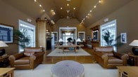 Located at 1000 Bally Bunion Drive in Egg Harbor, NJ, this single-story clubhouse offers a warm and well-appointed gathering space designed with both comfort and elegance in mind. The centerpiece of the interior is a grand open lounge area distinguished by soaring vaulted ceilings adorned with decorative string lighting and a ceiling fan, creating an airy yet intimate atmosphere. Rich hardwood flooring runs throughout, complementing the earthy tan wall tones and natural wood architectural accents. A classic fireplace flanked by built-in wooden shelving anchors one side of the room, adding a cozy, residential charm. The lounge is furnished with plush leather sofas and armchairs arranged around a wood coffee table atop a neutral area rug, providing an inviting setting for relaxation or casual conversation. Elegant table lamps cast a warm glow, enhancing the welcoming ambiance. Large windows along the perimeter flood the space with natural light and offer views of the surrounding greenery. A hallway visible through a central doorway connects to additional areas of the facility. On the opposite side, a countertop service area adds functional convenience to the space. Overall, the clubhouse exudes a refined yet approachable character, ideal for community gatherings and social events.