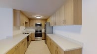 This single-floor space is highlighted by a thoughtfully designed kitchen that combines functionality with a clean, modern aesthetic. The kitchen features an abundance of light wood cabinetry that extends from floor to ceiling on both sides of the galley-style layout, providing generous storage throughout. The cabinetry is fitted with sleek bar-pull hardware, contributing to the contemporary feel of the space.

The countertops are finished in crisp white, offering a bright, clean work surface that contrasts beautifully with the warm wood tones of the cabinets. A full suite of stainless steel appliances includes an electric range with oven, an over-the-range microwave, and a French door refrigerator, all of which lend a polished, professional quality to the kitchen. A stainless steel sink is positioned beneath upper cabinetry on one side, completing the practical layout.

The flooring carries a light, neutral tone that complements the overall palette, and flush-mount ceiling lighting ensures the space is well-illuminated. The galley configuration maximizes efficiency while maintaining an open and airy ambiance throughout.