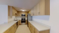 This single-floor space is highlighted by a thoughtfully designed kitchen that combines functionality with a clean, modern aesthetic. The kitchen features an abundance of light wood cabinetry that extends from floor to ceiling on both sides of the galley-style layout, providing generous storage throughout. The cabinetry is fitted with sleek bar-pull hardware, contributing to the contemporary feel of the space.

The countertops are finished in crisp white, offering a bright, clean work surface that contrasts beautifully with the warm wood tones of the cabinets. A full suite of stainless steel appliances includes an electric range with oven, an over-the-range microwave, and a French door refrigerator, all of which lend a polished, professional quality to the kitchen. A stainless steel sink is positioned beneath upper cabinetry on one side, completing the practical layout.

The flooring carries a light, neutral tone that complements the overall palette, and flush-mount ceiling lighting ensures the space is well-illuminated. The galley configuration maximizes efficiency while maintaining an open and airy ambiance throughout.