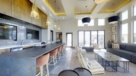 This elegantly designed single-floor space makes a striking impression with its soaring double-height ceilings and coffered ceiling details accented by warm ambient uplighting. The open-plan layout flows effortlessly from a stylish bar area to a generous lounge seating zone, creating a versatile environment suited for both social gatherings and relaxed conversation.

The bar counter, finished in a cool slate-toned surface, is lined with warm-toned leather barstools and flanked by rich, dark wood cabinetry housing a wall-mounted television — ideal for entertaining. A series of decorative gold pendant lights hang above the counter, adding a layer of glamour and warmth against the contemporary palette.

The lounge area features plush cream-toned seating arrangements paired with sleek, minimalist side tables, anchored by large-format floor tiles that unify the space. A bold drum pendant light serves as a focal centerpiece overhead.

Floor-to-ceiling glass doors flood the interior with natural light and open directly to an outdoor terrace or patio, seamlessly connecting the interior to the exterior environment. Windows positioned high on the walls further enhance the bright, airy atmosphere. The overall ambiance is modern and upscale, balancing comfort with sophisticated design details throughout.