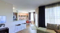 This single-floor space showcases an open-concept layout that seamlessly connects the kitchen, dining, and living areas. The kitchen features a granite-topped breakfast bar with sleek bar stools, providing both a functional prep surface and a casual seating option. Track lighting overhead illuminates the space efficiently, complementing the natural light that pours in through large windows dressed in floor-to-ceiling dark curtains, which add a dramatic contrast to the warm tones throughout. An accent wall painted in a soft peach hue adds warmth to the dining area, which is anchored by a compact dining table with seating. Open shelving in the background displays decorative greenery and accent pieces, contributing to the curated aesthetic of the space. Light hardwood-style flooring runs throughout, enhancing the sense of continuity between zones. A glass-paneled door leads to an outdoor area, further brightening the interior. The living area is partially visible, featuring a cushioned ottoman and neutral furnishings that complete the relaxed, contemporary atmosphere of this well-appointed space.