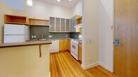Located at 210 Westminster Street in the heart of Providence, Rhode Island, this single-floor loft-style unit blends industrial character with modern functionality. The open-plan layout is anchored by expansive, light-toned hardwood floors that extend throughout the living areas, reflecting natural light that pours in through multiple double-hung windows lining the perimeter walls. Exposed white ductwork runs along the ceiling, lending an authentic industrial aesthetic, while a distinctive red structural column adds a striking architectural accent to the main living space.

The kitchen is thoughtfully designed with warm wood cabinetry, frosted glass upper cabinets with metal frames, and a sage green tile backsplash. Dark countertops provide generous prep space, and the kitchen opens to the living area via a breakfast bar with seating, creating a seamless connection between cooking and entertaining spaces.

The bathroom features neutral, large-format floor tiles, a full soaking tub with a tiled surround, and green mosaic tile accents, offering a clean and calm retreat. Track lighting and a ceiling fan enhance comfort and versatility throughout the unit. The row of windows overlooking the surrounding Providence streetscape fills the space with an abundance of natural light and urban energy, making this an ideal urban loft in a prime downtown location.