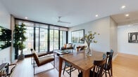 This well-designed single-floor residence showcases a thoughtful blend of modern aesthetics and functional living. The open-plan living and dining area is anchored by expansive floor-to-ceiling black-framed windows that flood the space with natural light and offer views of the surrounding streetscape and greenery. Light hardwood flooring runs throughout, complementing the neutral palette of the walls and ceiling. A ceiling fan adds a practical touch to the airy living space, which comfortably accommodates both a seating and dining arrangement.

The kitchen is outfitted with stainless steel appliances, high-gloss white lower cabinetry, natural wood upper cabinets, and a marble-effect countertop with under-cabinet lighting. The layout is efficient and clean, with recessed lighting enhancing the bright, modern feel.

The bedroom makes a bold design statement with deep forest green accent walls, large-format windows mirroring those in the living area, light hardwood floors, and a dramatic large-scale artwork above the bed. A gold tripod floor lamp and mirrored closet doors add refined detail.

The bathroom is finished in full marble-effect tile from floor to ceiling, featuring a glass-enclosed shower, a floating timber vanity with a black matte faucet, and a large mirror, creating a clean, spa-like atmosphere.