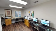 This single-floor business center is designed to provide a practical and efficient workspace for its users. The room is finished with durable wood-look flooring and neutral gray walls, creating a clean and professional atmosphere throughout. Overhead, flush-mounted fluorescent lighting ensures the space is well-lit for focused work at any time of day. A window fitted with white plantation shutters allows for natural light to filter in while maintaining privacy, adding a pleasant contrast to the otherwise utilitarian design. The workstation layout features dark granite-topped desks lining the walls, each equipped with desktop computers and ergonomic seating. A central wooden cabinet with a dark countertop serves as a functional island, housing a multifunction printer readily accessible to all users. A prominently displayed bulletin board on the wall provides a central location for notices, announcements, and community information. The space is thoughtfully organized to accommodate multiple users simultaneously, making it a convenient resource for printing, browsing, and completing work tasks. Overall, the business center combines a straightforward, no-frills design with practical amenities to deliver a reliable and comfortable environment for productivity.