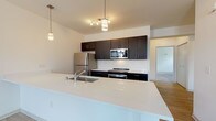 This single-floor unit offers a thoughtfully designed open-concept layout with modern finishes throughout. The kitchen serves as a central focal point, anchored by a large white quartz island with an integrated sink. Dark espresso cabinetry lines the walls, providing ample storage, while stainless steel appliances — including a refrigerator, microwave, and range — complete the contemporary look. Pendant lighting and a multi-bulb track fixture illuminate the space with warm light. Light wood-look flooring extends through the kitchen and common areas, transitioning to plush carpet in the bedroom.

The entryway features a warm wood front door with a transom window that draws in natural light. The bedroom is generously sized, with two windows offering outdoor views and filling the room with daylight. The bathrooms are finished with dark vanity cabinetry, white quartz countertops, and light wood-look flooring. One bathroom includes a walk-in shower and built-in open shelving for added storage. A dedicated laundry area houses a full-size washer and dryer alongside a water heater, adding practical convenience to this well-appointed space.