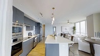 This single-floor space showcases a thoughtfully designed open-concept layout that seamlessly connects the kitchen and living areas. The kitchen is outfitted with sleek, matte grey cabinetry that extends to the ceiling, providing generous storage, while stainless steel appliances, including a French door refrigerator and a slide-in range, lend a professional touch. A long, white-topped kitchen island serves as the central gathering point, accommodating multiple bar-height metal chairs and offering additional workspace. Elegant pendant lights with brass hardware hang above the island, complementing the track lighting positioned over the cooking area. The open living space beyond features a ceiling fan with a light fixture, light wood-tone flooring throughout, and large windows that invite natural light into the room. A framed art piece anchors the living area wall, while lush indoor greenery adds a refreshing organic element. The overall aesthetic is clean, contemporary, and functional, with a neutral colour palette of whites and greys creating a cohesive and inviting atmosphere throughout.