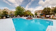 Located at 700 Mallards Way in O'Fallon, Missouri, this outdoor amenity area centers around a generously sized swimming pool designed for relaxation and recreation. The pool's most distinctive feature is a brick-based island planter positioned at its center, topped with a striking palm tree and accented with colorful flowering plants, creating a resort-inspired focal point. The surrounding pool deck is finished in smooth concrete and outfitted with numerous reclining lounge chairs and umbrella-shaded patio tables and chairs, offering ample space for sunbathing and casual outdoor gatherings. The perimeter is enclosed by a black iron fence, providing both security and an open, airy feel. Mature deciduous trees and vibrant seasonal plantings add natural beauty and shade throughout the area. A dedicated grilling station is also available, enhancing the outdoor entertaining experience. Adjacent to the pool, a charming clubhouse-style building with brick and siding detailing, large windows, and decorative flower boxes provides a welcoming backdrop. The multi-story residential buildings visible beyond the fence offer a sense of community. Overall, this well-maintained outdoor space delivers a comfortable, resort-like atmosphere suited for leisure and community enjoyment.