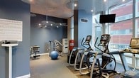 This modern fitness center is designed to provide a comprehensive workout experience within a sleek and inviting environment. The space is finished with cool blue-toned walls and a contemporary ceiling featuring distinctive cloud-shaped pendant light fixtures with recessed spotlights, creating a visually dynamic atmosphere. Large floor-to-ceiling windows along one wall flood the interior with natural light while offering views to the exterior, enhancing the open and energizing ambiance. The cardio zone is well-appointed with a row of elliptical trainers, each equipped with individual digital displays and entertainment screens, allowing members to track their progress during workouts. A wall-mounted flat-screen television is also positioned for easy viewing. The strength and functional training area includes a multi-station cable machine, an adjustable workout bench, and a stability ball, catering to a variety of exercise routines. A red Everlast punching bag adds a boxing and functional fitness element to the space. Storage solutions, including a small shelving unit, keep the area organized. The flooring transitions from a neutral carpet to rubberized matting in the equipment zones, providing appropriate support and safety. Overall, the fitness center combines practical functionality with modern design in a clean, well-lit setting.