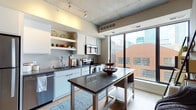 This single-floor urban unit showcases a distinctive industrial-modern aesthetic defined by exposed concrete ceilings throughout, which lend a raw, architectural character to the space. Track lighting runs along the ceiling, providing functional illumination while complementing the contemporary design. Light wood-tone flooring flows consistently across the open layout, adding warmth and visual continuity.

The kitchen is well-equipped with stainless steel appliances, including a refrigerator, range with microwave, and dishwasher. White cabinetry with open shelving lines the walls, and a small mosaic tile backsplash adds subtle texture. A freestanding kitchen table with a dark countertop serves as both a workspace and dining surface.

The living area flows directly from the kitchen and dining space, benefiting from expansive floor-to-ceiling windows that frame elevated city views and flood the room with natural light. A Murphy bed built into the wall offers a smart space-saving solution. The open floor plan creates a cohesive, multifunctional living environment.

The bathroom features a clean, minimalist design with a white vanity, integrated sink, ample under-counter storage, and a large mirror that enhances the sense of space. The overall ambiance is contemporary, efficient, and well-suited to urban living.