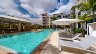 This outdoor amenity area centers around a generously sized swimming pool with vivid turquoise water, set against the backdrop of a multi-story Mediterranean-inspired residential building. The pool deck is finished in light-toned pavers, providing a clean and elegant surface that reflects the sunny atmosphere of the surroundings.

Along one side of the deck, a row of white lounge chairs is arranged beneath large white market umbrellas, offering shaded relaxation with direct views of the pool. On the opposite end, a covered cabana structure with a pitched metal roof provides an additional shaded gathering space, furnished for comfort and leisure.

Lush tropical landscaping, including tall palm trees and manicured hedges, frames the perimeter of the space, enhancing the resort-like ambiance. The multi-story building rising in the background features balconies that overlook the pool area, reinforcing the sense of an integrated, amenity-rich community. Open skies and abundant natural light contribute to the bright, welcoming character of this outdoor space, making it an ideal setting for relaxation and recreation.