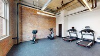 Located at 15 S Ontario St in downtown Toledo, Ohio, this fitness room offers a well-appointed exercise space that blends industrial character with modern functionality. The room is defined by its striking exposed brick accent wall, which runs the length of the space and adds a warm, textural contrast to the crisp white walls on the opposing side. Above, an open ceiling reveals raw concrete, visible ductwork, and exposed piping — a nod to the building's historic industrial roots — while linear LED lighting ensures the space is well-lit throughout.

The fitness room is outfitted with cardio equipment including treadmills, stationary bikes, and an elliptical machine, all positioned on durable rubber flooring designed for high-impact use. A large window along the brick wall allows natural light to filter in, offering views of the surrounding downtown streetscape. The space is accessed through a full-height glass wall and door system, providing a transparent, open visual connection to the adjacent corridor or lobby area.

A concrete support column anchors the open floor plan, while a dedicated door on the brick wall side provides additional access or utility. The overall ambiance is modern and energetic, combining the raw aesthetic of the building's architecture with clean, purpose-driven fitness amenities.