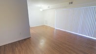 This single-floor space offers a clean, open living area defined by smooth, warm-toned hardwood-style flooring that extends throughout the room, reflecting light and contributing to the overall brightness of the interior. The walls are finished in crisp white, creating a neutral and versatile backdrop well-suited to a variety of furnishing styles. A large window fitted with vertical blinds dominates one wall, allowing for adjustable natural light and lending the space an airy, open quality. The ceiling is standard height and features a central light fixture, complemented by ceiling-mounted HVAC vents that suggest a centralized climate control system. The layout appears open and unobstructed, providing flexibility in how the space can be arranged and utilized. Baseboards run along the perimeter of the room, adding a clean, finished look to the flooring transitions. Overall, the space presents a well-maintained, minimalist environment with practical finishes that balance functionality and aesthetic simplicity, making it a adaptable setting ready for occupancy.