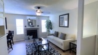 The living room serves as the central gathering area of this single-story space, designed with a clean, neutral palette that creates a warm and inviting atmosphere. A brick-surround fireplace anchors one wall, topped with a decorative mirror and accented by a sunburst wall ornament, adding a touch of character to the room. A ceiling fan with a built-in light fixture provides both functionality and soft illumination overhead, complemented by a textured popcorn ceiling.

Natural light filters in through multiple windows, brightening the space and highlighting the hardwood-style flooring beneath. A patterned area rug in dark tones grounds the seating arrangement, which features a neutral-toned sofa adorned with coordinating pillows. A glass-top coffee table with a dark metal frame sits at the center, offering both style and practicality. A tall indoor plant introduces a refreshing natural element, while framed wall art adds a finishing decorative touch. The overall ambiance is comfortable, tidy, and well-appointed.