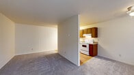 This well-organized one-bedroom, one-bathroom unit offers an efficient single-floor layout that maximizes every square foot. The combined kitchen and living area forms the heart of the home, with the two spaces separated by a partial wall partition. The kitchen features warm-toned wood cabinetry, granite-style countertops, hardwood flooring, and white appliances including an electric range and refrigerator, with a peninsula providing additional counter space and a natural boundary between cooking and living areas. The living room is generously sized with soft carpet flooring, a ceiling fan, and large windows alongside the front entry door that allow abundant natural light and outdoor views. A wall-mounted heater unit provides functional climate control. The bedroom is a comfortable, carpeted retreat featuring a wide window with blinds, a wall heater, and an open closet with built-in shelving for storage. The bathroom is clean and bright, appointed with white tile surrounding a full soaking tub and shower combination, complemented by a small window for ventilation. A hallway connects the main living spaces to the bedroom and bathroom, completing this straightforward and practical floor plan.
