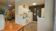 This single-floor space offers a thoughtfully designed open-concept layout that seamlessly connects the kitchen and dining areas. The kitchen is well-appointed with crisp white cabinetry, white appliances including a gas range and full-size refrigerator, and a neutral countertop that complements the overall color palette. A bold dark accent wall adds a striking visual contrast to the otherwise light and airy interior, creating a focal point that adds character and depth to the space. Decorative oversized utensil wall art reinforces the kitchen's identity and adds a playful yet intentional design touch. The warm wood-style flooring runs throughout, providing a cohesive and inviting foundation underfoot. A patterned area rug in the kitchen zone adds texture and definition to the space. The dining area is adjacent, accommodating a wooden dining table that ties naturally into the warm tones of the flooring. Soft, ambient ceiling lighting illuminates the space evenly. An open passageway connects to a secondary living area visible in the background, enhancing the sense of flow and continuity throughout the home.