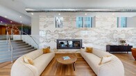 This expansive single-floor space is designed with a sophisticated, contemporary aesthetic, featuring warm wood flooring throughout that unifies its numerous distinct zones. The centerpiece of the main lounge is a floor-to-ceiling bookmatched marble accent wall with striking gold and grey veining, housing a sleek double-sided fireplace flanked by paired cream sofas and accent chairs. Exposed metallic ductwork above adds an industrial contrast to the polished marble surface, while clerestory windows allow views to the exterior cityscape beyond.

A grand curved staircase with stainless steel railings anchors one corner of the space, adjacent to a dramatic large-scale water feature set against a textured dark stone wall, creating a calming focal point. The main entry area features a soaring rotunda-style ceiling with exposed brick columns, abundant natural light from large perimeter windows, and generous open seating arrangements with tropical plant accents.

Additional lounge areas include a reading nook with built-in light wood shelving, a leather seating zone, a flexible lounge with modular grey furniture, and a casual area with mixed seating arrangements. Recessed lighting throughout ensures a consistent, welcoming ambiance across every zone.