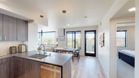 This contemporary single-floor residence showcases a thoughtfully designed open-concept layout that seamlessly connects the kitchen, dining, and living areas. The kitchen is a standout feature, equipped with dark wood-grain cabinetry, a tile backsplash, stainless steel appliances including a French door refrigerator and range, and a spacious center island with a quartz countertop that doubles as a breakfast bar with seating. Light hardwood flooring runs throughout the main living areas, complementing the clean white walls and modern recessed lighting.

The dining area comfortably accommodates a full-sized table and is framed by large black-framed windows and a glass door that opens to a balcony, offering treetop and neighborhood views. The living room benefits from an oversized picture window that floods the space with natural light and continues those scenic outdoor views.

The residence includes two well-proportioned bedrooms, each carpeted and featuring black-framed windows with views of the surrounding neighborhood. The bathrooms are finished in a clean, minimalist style with white cabinetry, quartz countertops, large mirrors, and neutral tile flooring. Overall, the space presents a cohesive modern aesthetic with an emphasis on natural light and open, functional living.