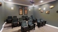 This private home theater is thoughtfully designed to replicate the ambiance of a traditional cinema. The room is finished with deep grey walls accented by crisp crown molding, creating a refined yet intimate atmosphere. Rows of plush, dark upholstered theater-style seats with fold-down cushions are arranged to ensure clear sightlines for all viewers, accommodating a generous number of guests comfortably. A ceiling-mounted projector anchors the technical setup, positioned to deliver a high-quality image to the screen. The patterned carpet underfoot adds a classic movie-house aesthetic while contributing to the room's acoustics. Wall-mounted sconce lighting provides warm, subtle illumination, enhancing the cinematic ambiance without disrupting the viewing experience. Decorative vintage-style cinema and theatre posters in ornate frames adorn the walls, reinforcing the space's dedicated entertainment purpose. In-wall speakers are also visible, suggesting a well-integrated surround sound system. Overall, this screening room offers a polished, fully immersive entertainment environment ideal for private film screenings and events.