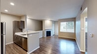 Located at 9113 W Cross Dr in Littleton, Colorado, this single-floor residence offers a thoughtfully designed open-concept layout that seamlessly connects the kitchen, dining, and living areas. Rich hardwood-style flooring flows throughout the main living spaces, complemented by neutral warm-toned walls and clean white trim that create a bright, welcoming ambiance.

The kitchen is well-appointed with white upper cabinetry, a stainless steel refrigerator, microwave, and range, alongside a peninsula with a light countertop that provides both prep space and a casual seating area. A stainless steel sink with recessed lighting above adds a polished, functional touch.

The living area is anchored by a fireplace with a crisp white mantle, making it an inviting focal point. A large window allows abundant natural light to fill the room and offers views of the surrounding neighborhood. An additional room, finished with plush carpeting and a ceiling fan, serves as a comfortable bedroom with its own window for natural light.

The bathroom is cleanly finished with wood-look flooring, a tiled tub-shower combination, dark-toned vanity cabinetry, and brushed nickel fixtures. A staircase visible in the model view suggests access to additional levels or a lower entry. Overall, the space presents a clean, modern aesthetic well-suited for comfortable everyday living.