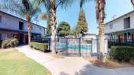 This single-story residential community presents a welcoming Southern California aesthetic, characterized by mature palm trees, verdant lawns, and thoughtfully maintained landscaping throughout its grounds. The exterior architecture features stucco construction with a neutral gray finish, complemented by bold red accent doors that add a distinct visual character to the building façade.

The community is fully enclosed by a robust black iron perimeter fence anchored by substantial stone masonry pillars, providing residents with a secure and private environment. Access is controlled through a gated entry, reinforcing the community's commitment to safety and exclusivity.

A standout amenity is the community swimming pool, enclosed within its own dedicated black iron fence and self-closing gate. The pool area is framed by swaying palm trees and manicured hedges, creating a resort-like atmosphere ideal for relaxation. Concrete walkways wind gracefully throughout the property, connecting residents to the pool and other common areas.

The grounds are generously landscaped with a mix of tall palms, broad shade trees, trimmed shrubs, and patches of green lawn, contributing to a vibrant and inviting outdoor atmosphere. Clear blue skies overhead reinforce the warm, sun-drenched climate characteristic of the Southern California region, making the outdoor spaces a natural extension of daily living.