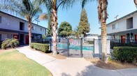 This single-story residential community presents a welcoming Southern California aesthetic, characterized by mature palm trees, verdant lawns, and thoughtfully maintained landscaping throughout its grounds. The exterior architecture features stucco construction with a neutral gray finish, complemented by bold red accent doors that add a distinct visual character to the building façade.

The community is fully enclosed by a robust black iron perimeter fence anchored by substantial stone masonry pillars, providing residents with a secure and private environment. Access is controlled through a gated entry, reinforcing the community's commitment to safety and exclusivity.

A standout amenity is the community swimming pool, enclosed within its own dedicated black iron fence and self-closing gate. The pool area is framed by swaying palm trees and manicured hedges, creating a resort-like atmosphere ideal for relaxation. Concrete walkways wind gracefully throughout the property, connecting residents to the pool and other common areas.

The grounds are generously landscaped with a mix of tall palms, broad shade trees, trimmed shrubs, and patches of green lawn, contributing to a vibrant and inviting outdoor atmosphere. Clear blue skies overhead reinforce the warm, sun-drenched climate characteristic of the Southern California region, making the outdoor spaces a natural extension of daily living.