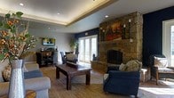 This inviting single-floor space makes an immediate impression with its thoughtfully designed living area, where comfort and character come together seamlessly. The focal point of the room is a commanding floor-to-ceiling stone fireplace, crafted from natural stacked stone and topped with a rustic mantel adorned with candle accents and a vibrant piece of artwork. Flanking the fireplace, plush navy blue seating and a solid wood coffee table rest atop a woven area rug, creating a cozy and cohesive gathering space. A tray ceiling with recessed lighting adds architectural depth and a refined touch to the room, while a bold navy blue accent wall grounds the space with a sense of sophistication. French doors along the back wall allow generous amounts of natural light to flood the interior, offering a bright contrast to the warm, earthy tones throughout. A casual dining or lounge area with wicker-style chairs and a wall-mounted television is visible in the background, adding to the versatility of the layout. Lush indoor greenery contributes a natural, refreshing element to the overall ambiance. The space strikes a balance between relaxed and refined, making it well-suited for both casual gatherings and comfortable everyday use.