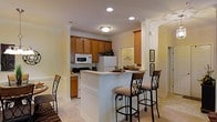 Located at 901 Falls Park Dr in Sanford, NC, this single-story residence offers a thoughtfully designed interior with a warm and inviting atmosphere throughout. The kitchen serves as a central highlight of the home, showcasing rich medium-toned wood cabinetry that extends to the upper walls, providing generous storage space. Crown molding adds a refined architectural touch, framing both the cabinetry and the ceiling with elegant detail. The kitchen flows seamlessly around a peninsula-style breakfast bar, accented by decorative columns and topped with a light countertop surface. Cushioned bar stools with ornate wrought iron frames line the bar, creating a casual dining and gathering spot. Recessed lighting and a flush-mount ceiling fixture illuminate the space efficiently, complementing the light-toned tile flooring that runs throughout. Full appliances, including a range, refrigerator, and built-in microwave, are integrated into the cabinetry. An adjacent area, visible through a pass-through opening, hints at additional living or utility space. The overall palette of soft cream walls, warm wood tones, and light flooring creates a cohesive, comfortable environment well-suited for everyday living.