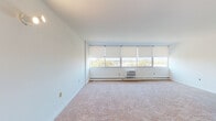 Located at 2400 McClellan Ave in Pennsauken, NJ, this single-floor unit offers a clean, functional layout with well-defined living spaces throughout. The generously sized living area is a standout feature, characterized by a wide bank of floor-to-near-ceiling windows fitted with roller shades that flood the room with natural light while offering elevated views of the surrounding landscape and treetops. Wall-to-wall carpeting in a neutral tone runs throughout the living and bedroom areas, contributing to a quiet, comfortable atmosphere. A wall-mounted air conditioning unit and baseboard heating provide year-round climate control. The bedroom is a well-proportioned space with built-in closet storage and soft wall sconce lighting, maintaining the same clean, understated aesthetic found throughout the unit. The galley-style kitchen is efficiently designed, featuring warm oak cabinetry on both sides, generous counter space, a gas range, refrigerator, and a stainless steel sink, all set on durable tile flooring. The bathroom is neatly appointed with white cabinetry, a dark-toned countertop with an undermount sink, a medicine cabinet with mirror, tile flooring, and a tub-shower combination, delivering a practical and tidy finish. Overall, the unit presents a straightforward, well-maintained living space with ample natural light and functional amenities.