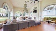 The Veranda Area at The Residences at Bella Terra is a sophisticated semi-outdoor amenity space defined by its striking Mediterranean-inspired architecture. Soaring ceilings, grand arched openings adorned with decorative ironwork fanlight details, and slender classical columns frame panoramic views of a lush, manicured courtyard lawn surrounded by mature trees and well-maintained hedging.

The main lounge area is anchored by a tall white fireplace with a mounted flat-screen television above it, creating a comfortable focal point for gatherings. Plush sectional sofas, accent chairs, and a low coffee table are arranged on a large area rug, while woven globe pendant lights and a ceiling fan illuminate the space. A substantial solid-wood dining table with wicker chairs provides ample seating for communal meals or activities.

An adjacent outdoor kitchen is equipped with stainless steel appliances, generous stone-topped countertops in an L-shaped configuration, and ample cabinet storage — ideal for entertaining. Intimate seating nooks positioned beneath the arches offer quieter retreats with views of the courtyard's greenery. Throughout, warm terracotta-toned flooring ties the space together, contributing to the veranda's inviting and refined ambiance.