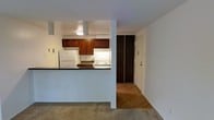 This single-floor space showcases a practical and open layout, with the kitchen and living area flowing seamlessly into one another. The kitchen is defined by rich, warm-toned wood cabinetry that provides ample storage, complemented by dark countertops that offer a subtle contrast to the white walls and appliances. A white refrigerator and electric range contribute to a clean, functional cooking space, while under-cabinet lighting enhances visibility and adds a welcoming glow. A half-wall peninsula separates the kitchen from the adjacent living area, creating a natural division of space while maintaining an open feel. The living area features neutral-toned carpet, providing a comfortable underfoot experience, while wood-look flooring transitions into the hallway beyond. A sliding door closet with dark wood-paneled doors adds storage convenience near the entryway. The walls are painted in a clean, bright white throughout, contributing to an airy atmosphere. The straightforward, single-level layout ensures easy navigation between spaces, making this unit well-suited for those seeking a low-maintenance, efficiently designed living environment.