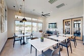 Located at 5801 S 51st Ave in Phoenix, AZ, this single-floor community gathering space combines a dining area and a fully equipped kitchen into one cohesive, modern environment. The dining area is bright and airy, featuring large floor-to-ceiling sliding glass doors and clerestory windows that flood the space with natural light and offer views of an outdoor patio. Multiple white marble-top dining tables paired with sleek black chairs create a clean, contemporary aesthetic, while bold pendant lights with black-and-gold finishes add visual interest. A large ceiling fan with oversized blades provides comfort overhead. A lounge area with neutral-toned seating and a wall-mounted television anchors the back of the room, complemented by decorative wall art and sculptural 3D wall panels. The kitchen is equally refined, boasting dark charcoal cabinetry with brass hardware, a white subway tile backsplash, open wood floating shelves, and stainless steel appliances. A large white island with built-in seating for multiple guests serves as the focal point, illuminated by a striking globe chandelier in brushed gold. Light tile flooring and abundant window coverage throughout unify the space, creating a welcoming and functional environment ideal for communal dining and gathering.