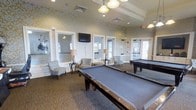 This single-floor recreational space is thoughtfully designed to offer both comfort and entertainment. The room is anchored by two full-sized billiard tables with blue felt surfaces, providing ample space for gameplay and social interaction. Decorative pendant lighting hangs from the ceiling, complementing the recessed lights and casting a warm, inviting glow throughout the space.

The walls are adorned with an elegant gold and cream damask-patterned wallpaper, adding a refined aesthetic to the room. White wainscoting and trim details contribute a classic architectural character. Large windows line the walls, allowing natural light to filter in and offering views of the surrounding exterior.

Seating areas are arranged thoughtfully around the perimeter, featuring upholstered accent chairs paired with side tables and table lamps, creating comfortable spots for guests to relax and socialize. A wall-mounted flat-screen television provides additional entertainment options.

The space features a tray ceiling detail that adds architectural depth and visual interest. A glass-paneled door at the rear of the room provides access to an adjacent area or exterior. The overall ambiance blends a classic, elegant design with a welcoming, leisure-focused atmosphere, making it well-suited as a community or clubhouse amenity.