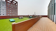 Located atop 855 Main St in downtown Bridgeport, Connecticut, this rooftop amenity level offers a well-appointed outdoor retreat with sweeping views of the city skyline and surrounding urban landscape. The space is divided into two distinct functional zones, each designed to maximize usability and comfort.

The first zone features a lush artificial turf area enclosed by warm wood-paneled and brick perimeter walls. This dedicated pet-friendly area is equipped with agility structures, including tunnel and shelter elements, making it an ideal space for dogs to play and exercise. A gated entry provides secure access to this section.

The second zone is an expansive composite wood deck finished in a rich cedar tone. Multiple seating arrangements, consisting of modern teak-framed chairs and sofas with plush cream cushions, are thoughtfully positioned throughout the deck, offering flexible gathering spaces for residents. A large glass curtain wall facade serves as a striking architectural backdrop to this lounge area, reflecting the surrounding cityscape.

Together, both areas are bordered by classic brick parapet walls that frame panoramic views of Bridgeport's downtown buildings and open horizon. The rooftop's open-air design ensures ample natural light throughout the day, creating an inviting urban escape above the city streets.
