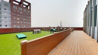 Located atop 855 Main St in downtown Bridgeport, Connecticut, this rooftop amenity level offers a well-appointed outdoor retreat with sweeping views of the city skyline and surrounding urban landscape. The space is divided into two distinct functional zones, each designed to maximize usability and comfort.

The first zone features a lush artificial turf area enclosed by warm wood-paneled and brick perimeter walls. This dedicated pet-friendly area is equipped with agility structures, including tunnel and shelter elements, making it an ideal space for dogs to play and exercise. A gated entry provides secure access to this section.

The second zone is an expansive composite wood deck finished in a rich cedar tone. Multiple seating arrangements, consisting of modern teak-framed chairs and sofas with plush cream cushions, are thoughtfully positioned throughout the deck, offering flexible gathering spaces for residents. A large glass curtain wall facade serves as a striking architectural backdrop to this lounge area, reflecting the surrounding cityscape.

Together, both areas are bordered by classic brick parapet walls that frame panoramic views of Bridgeport's downtown buildings and open horizon. The rooftop's open-air design ensures ample natural light throughout the day, creating an inviting urban escape above the city streets.