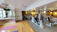 This fitness center is thoughtfully designed to accommodate a variety of workout routines within a single, well-organized space. The room is divided into distinct functional zones, with polished hardwood flooring in the stretching and yoga area, where colorful exercise mats are laid out for floor-based workouts. This transitions seamlessly into a carpeted section housing an array of cardio equipment, including treadmills and an upright exercise bike, ideal for endurance training. A multi-station strength training machine anchors the space, offering users a comprehensive resistance workout. Full-length mirrors line portions of the walls, enhancing the sense of openness and allowing for form checks during exercise. The interior is finished with elegant white crown molding and warm wall sconces set against rich, earth-toned accent walls, giving the space a refined yet functional character. Overhead lighting complements the ambient wall fixtures, ensuring the room is well-lit throughout. The overall design strikes a balance between practicality and aesthetic polish, making this fitness center a comfortable and fully equipped environment for a wide range of physical activities.