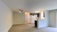 This single-floor space showcases an open-concept design that seamlessly connects the living area and kitchen, creating a cohesive and functional layout. The living area is finished with wall-to-wall carpeting in a neutral beige tone, complemented by light gray walls and white baseboards that lend a clean, contemporary feel. A ceiling fan with a light fixture is centrally positioned, providing both airflow and ambient lighting throughout the space.

The kitchen is well-appointed with dark espresso cabinetry that provides a strong contrast against the white appliances, including a refrigerator, microwave, and range. A peninsula with a light-colored countertop extends into the living space, offering additional prep area and serving as a natural divider between the two zones. Recessed lighting and a flush-mount ceiling fixture ensure the kitchen is well-lit. A window above the sink allows for natural light to filter in. A hallway visible from the main living area leads to additional rooms, suggesting a practical and straightforward floor plan throughout the unit.