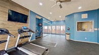 This single-floor fitness center is designed to support a wide range of workout routines in a bright, energizing environment. The space is defined by bold blue walls and durable dark rubber flooring, creating a dynamic atmosphere conducive to focused exercise. Ceiling fans are positioned throughout, ensuring consistent airflow and comfort during workouts.

The cardio zone is anchored by multiple treadmills and an elliptical machine, each equipped with individual display screens. A wall-mounted flat-screen television is set against a wood-panel accent wall, providing entertainment during cardio sessions. Adjacent to the cardio area, a dedicated stretching and open-floor zone offers ample room for mat work, yoga, or group exercise.

A full-length barre rail runs along a wall-to-wall mirror, making the space suitable for barre, dance, or flexibility training. The mirrored wall also enhances the sense of spaciousness throughout the room. A rack of weighted balls and additional accessories provides options for strength and functional fitness training.

Large windows along one wall invite generous natural light and offer views of the surrounding outdoor area. A service window built into one wall suggests access to an adjacent support area. Multiple entry points, including glass double doors, connect the fitness center to the exterior, further enhancing ventilation and accessibility.