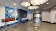 This lobby presents a polished, contemporary aesthetic that sets a professional and welcoming tone. The space is defined by large-format tile flooring in a warm neutral tone, which reflects both natural and artificial light to enhance the sense of openness. Oversized drum pendant light fixtures suspended from the ceiling serve as a striking focal point, providing generous illumination while adding a bold design statement overhead. Recessed lighting throughout the ceiling further brightens the space evenly.

A prominent dark tile accent wall creates a strong visual anchor, offering contrast against the lighter surrounding walls and lending depth to the interior. A sleek console table with a wood-and-metal frame is positioned along one wall, accented by a vibrant abstract artwork and a white orchid arrangement, adding a touch of color and organic warmth. A compact seating area nearby features upholstered chairs in a neutral pattern, ideal for brief gatherings or waiting.

The corridor extends toward a set of glass-paneled entry doors at the far end, allowing natural light to filter into the space. Overall, the lobby combines functional design with refined finishes to create an inviting and sophisticated environment.