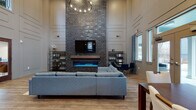 Located at 22000 NE Halsey St in Fairview, Oregon, this single-story community space is defined by its striking architectural scale and modern design. The main gathering area commands attention with dramatically tall ceilings that rise well above two stories in height, accentuated by clerestory windows positioned near the top of the walls. These upper-level windows, combined with a bank of large windows and French doors along the exterior wall, flood the interior with abundant natural light and provide views of the surrounding outdoor area.

A floor-to-ceiling dark brick accent wall serves as the room's focal point, anchoring a contemporary linear electric fireplace with a wood mantel shelf and a wall-mounted television above. The open layout accommodates a generously sized sectional sofa and coffee table as the central lounge seating, complemented by a secondary seating nook with a glass-top table and chairs. A dedicated dining area with a dark wood table and upholstered seating is positioned near the French doors.

Warm-toned luxury vinyl plank flooring runs throughout the main space. A separate kitchenette offers dark cabinetry, light countertops, a sink, stainless steel appliances including a refrigerator and dishwasher, and dark tile flooring, providing practical amenities for gatherings.